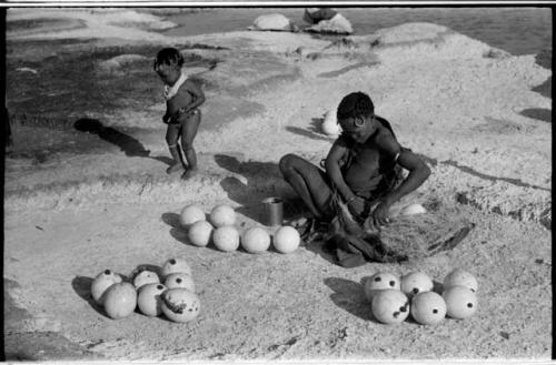 Khuan//a (Gau's wife) placing ostrich egg shells wrapped in grass in her kaross and her child standing near her