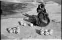 Khuan//a (Gau's wife) placing ostrich egg shells wrapped in grass in her kaross and her child standing near her