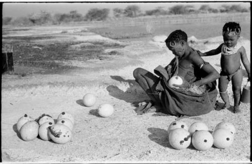 Khuan//a (Gau's wife) placing ostrich egg shells wrapped in grass in her kaross with her child standing beside her