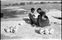 Khuan//a (Gau's wife) placing ostrich egg shells into her kaross with her child standing beside her