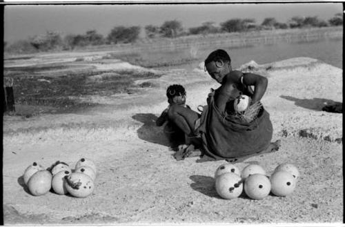 Khuan//a (Gau's wife) placing ostrich egg shells into her kaross with her child crouching beside her