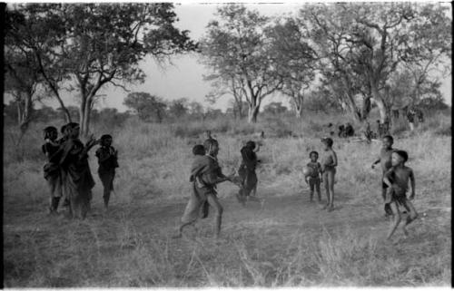 Girls playing N!owa t/ama (melon tossing game) / tamah n!o’an (ball game); four girls to the left are singing and clapping and an unidentified, visiting girl in the center carries a child on her back while she plays; other children present in the background