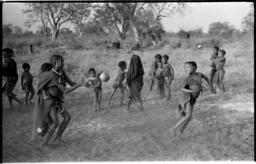 Girls playing N!owa t/ama (melon tossing game) / tamah n!o’an (ball game); an unidentified, visiting girl carries a child on her back as she plays; other children present in the background