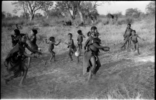 Girls playing N!owa t/ama (melon tossing game) / tamah n!o’an (ball game); an unidentified, visiting girl carrying a child on her back tosses the melon as !Ungka's cousin runs to catch it; other children present in the background