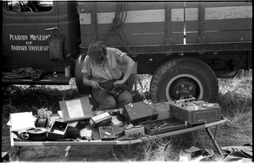 Elizabeth Marshall Thomas sitting in front of the expedition truck monitoring the recorder with equipment laid out in front of her