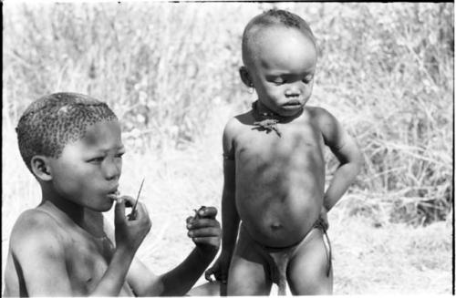 /Gaiamakwe eating a piece of meat, with N!whakwe standing next to him