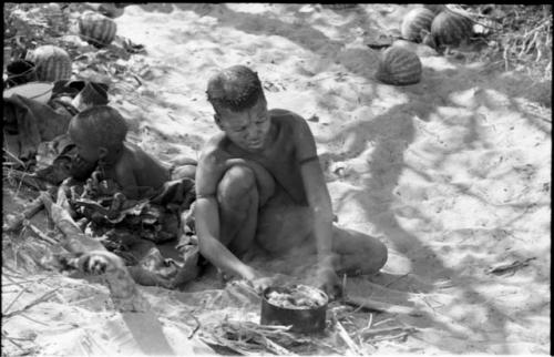 Tsekue cooking a stew of meat and tsama melon pulp at the fire, with N!whakwe beside her