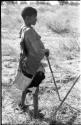 DaSi!Na in profile standing and leaning against digging stick, showing apron around her waist