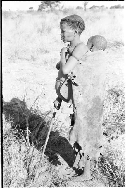 Tsekue in a kaross with N!whakwe tied to her back, standing holding a digging stick