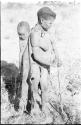 Tsekue wearing a kaross, with N!whakwe tied to her back, holding a digging stick, full figure