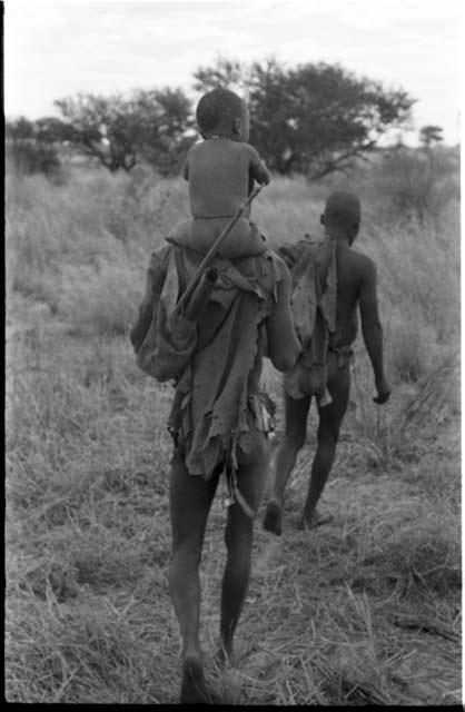 Man carrying a child on his shoulders; with another man walking ahead of him, seen from behind