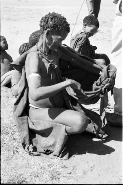 Woman holding out her kaross for tobacco, with other people in the background