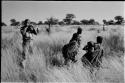 /Twikwe, DaSi!Na, and Tsekue carrying N!whakwe, walking in grass; Bill Donnelan taking a photo