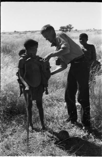 Tsekue, with N!whakwe on her back, standing beside John Marshall, DaSi!Na in the background