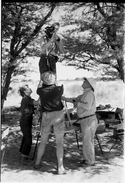 John Marshall standing on a table, holding a film camera; Lorna Marshall, Laurence Marshall, and an expedition member holding camera tripod steady