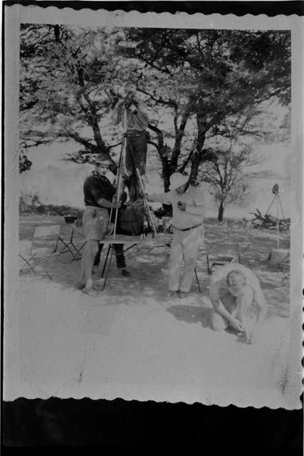 John Marshall standing on a table with a camera on a tripod, Laurence Marshall and an expedition member holding the tripod; another expedition member kneeling