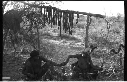 ǂToma and //Kushay sitting by a fire, beneath a trestle with meat hanging from it