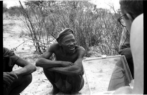 Wilhelm Camm and Nicholas England in chairs with a sound recorder interviewing two men sitting on the ground, close-up
