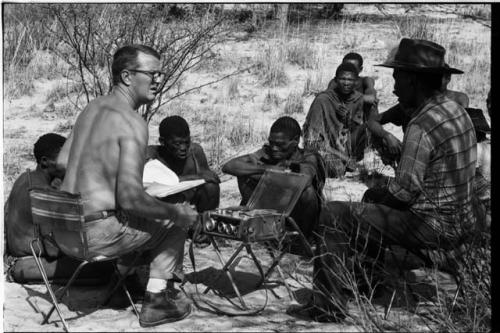 Wilhelm Camm and Nicholas England in chairs with a sound recorder interviewing a group of men sitting on the ground