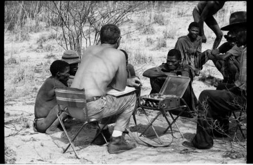 Wilhelm Camm and Nicholas England in chairs with a sound recording machine interviewing a group of men sitting with them