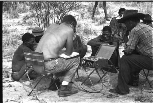 Wilhelm Camm and Nicholas England in chairs with a sound recording machine interviewing a group of men sitting with them