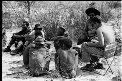 Wilhelm Camm and Nicholas England sitting in chairs with a sound recording machine taking notes, interviewing a group of men sitting on the ground with them