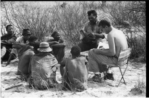 Wilhelm Camm and Nicholas England sitting in chairs with a sound recording machine taking notes, interviewing a group of men sitting on the ground