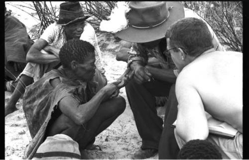 Man squatting, pointing to his palm, being interviewed by Wilhelm Camm and Nicholas England; two people in the background