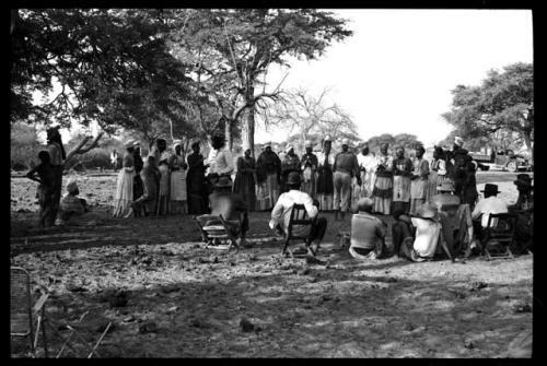 Group of women clapping, standing in a line with Judas Kangengi standing in the middle, preparing to sing; people watching