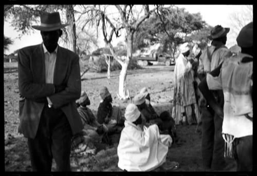 Man standing with his arms crossed; a group of women sitting and a group of people standing in the background