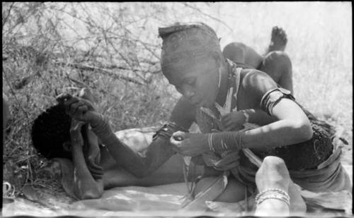 Woman holding a thread, with a baby on her back in her kaross, and a person lying down behind her