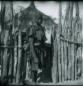 Woman carrying a baby on her back, standing in front of fence with pole tied with creepers