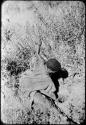 Woman digging, her digging stick in her left hand, pulling sand out of a hole with her right hand
