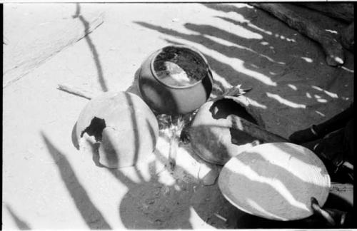 Two broken pots, one whole pot, and a basket holding grain meal; a woman's hand is holding a pestle and the basket