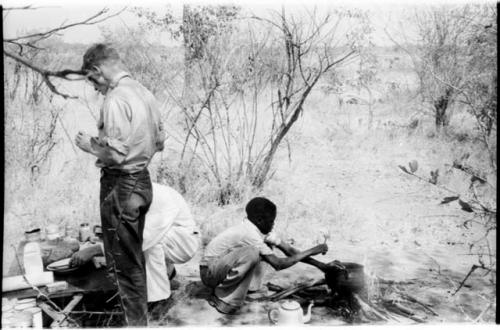 Two men at the expedition camp and a boy crouched over a pot on a fire