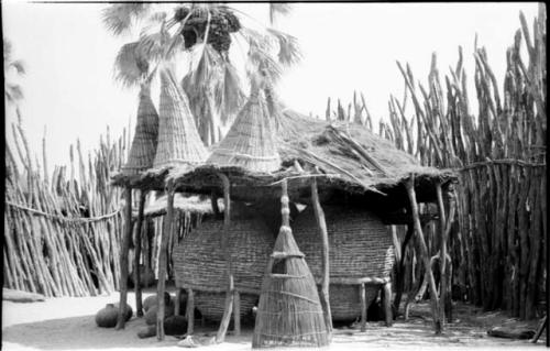 Large storage baskets supported by poles under a thatched roof in an Ombandia kraal; several fish baskets are on the roof and one is on the ground; palm tree in the background