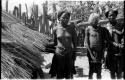 Three girls standing by a hut; one of the girls wears a beaded headdress