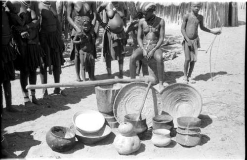 Pots, baskets, and gourds in the foreground; people in the background including a seated girl and a boy with a bow and arrow