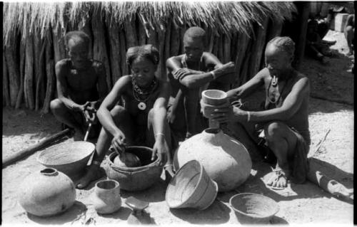 Two women and two men, sitting among gourds and grain baskets; one woman holds a funnel over the mouth of a gourd while the other woman scoops meal with a gourd ladle