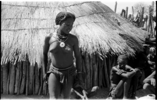 Girl standing beside a hut; other people seated in the background