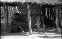 Man sitting under a thatched roof next to large storage basket supported by wooden struts