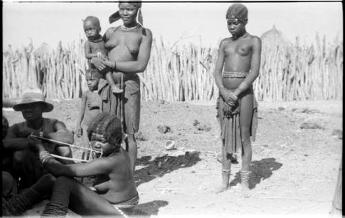 Girl sitting and playing a musical bow and a man wearing a hat in the foreground; two girls and a child standing in the background, one girl holds a baby