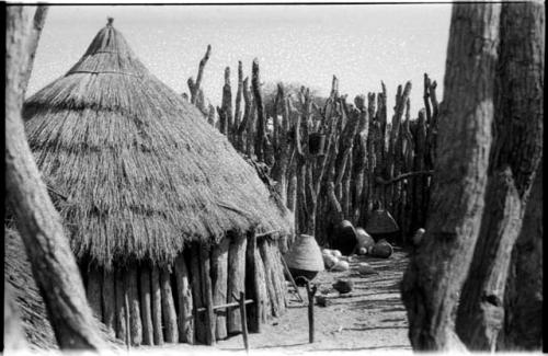 Hut with a door locked by a pole; baskets and pots by fence in the background