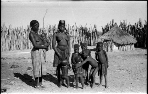 Two women and seven children, one held by a woman; wood pole fence in the background