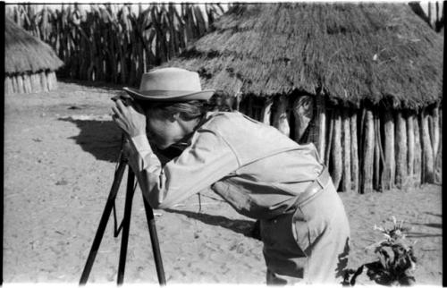 Lorna Marshall using a camera on a tripod; hut and wood pole fence in the background