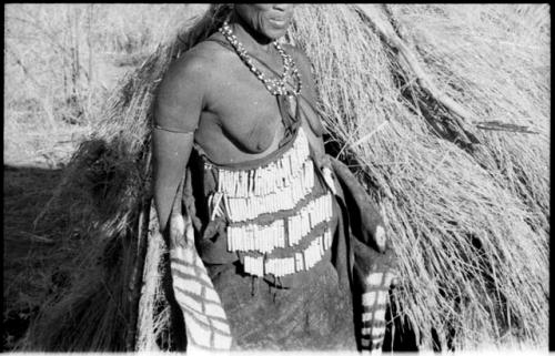 Woman standing, wearing an ornamented kaross and beaded apron, partial figure