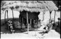 Storage basket supported by wooden struts surrounded by pots, all under a thatched roof