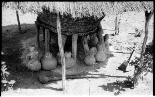 Storage basket supported by wooden struts surrounded by pots, all under a thatched roof