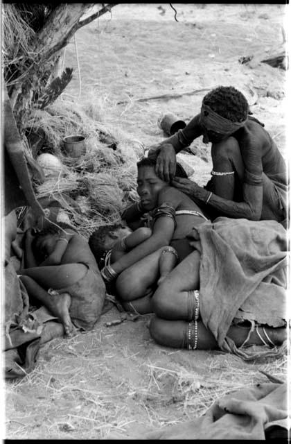 Elderly woman adjusting the earring of a younger woman who is lying with her baby; another child asleep beside them