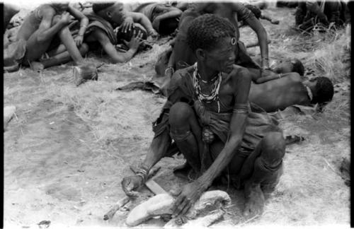 "Old Xama" preparing a piece of animal skin, other unidentified people in the background